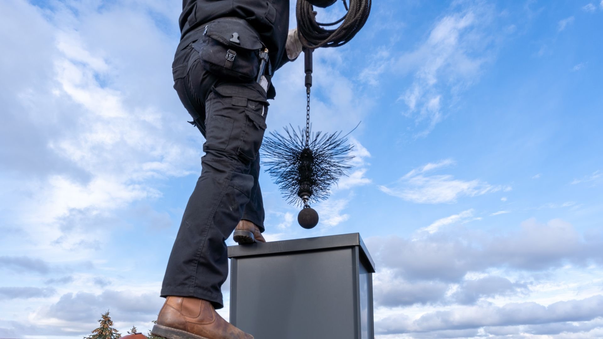 Chimney sweep standing on box, holding brushing tool against blue sky